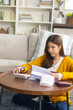 © kan - A focused young Asian woman sits at a desk in her living room at home, using a calculator to plan her monthly budget plan, ensure her bills, and focus on managing her expenses and financial goals.