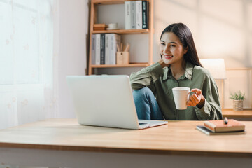 Wall Mural - young woman sits at wooden desk with laptop, holding coffee mug and smiling, enjoying relaxed moment in bright home office with bookshelves and natural light