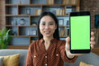 © Liubomir - Asian woman in casual attire shows smartphone with vibrant green screen at home. Focus on phone that is held towards camera, inviting concepts of technology, communication, apps.