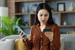 © Liubomir - Asian woman at home looking worried holding smartphone and credit card, symbolizing online shopping, financial issues, or mobile banking problem. Home interior setting adds to domestic feel.