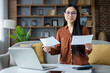 © Liubomir - Asian woman with glasses smiling while reviewing documents at home office with laptop. Comfortable, professional setting, perfect for remote work or studying.