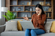 © Liubomir - Asian woman feeling stressed while working on laptop at home, sitting on cozy couch surrounded by cushions and plants. Uncertain expression indicates work-from-home challenges.
