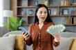 © Liubomir - Excited woman holds cash while checking her phone, indicating online earnings success from home. Asian woman sits on cozy sofa in modern living room, expressing financial achievement and happiness.