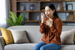 © Liubomir - Asian woman using asthma inhaler, sitting on sofa experiencing breathing issue. Wearing polka dot blouse, managing respiratory health at home. Focused, calming atmosphere.