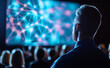 © Curioso.Photography - A speaker presenting a glowing blue network visualization on a large screen to an audience, symbolizing AI, machine learning, and connectivity.