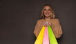 © My Ocean studio - A cheerful woman in a beige sweater smiles brightly, holding vibrant yellow, pink, and green shopping bags against a dark background, exuding joy and enthusiasm for shopping and retail therapy.