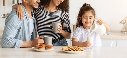 Wall Mural - Loving middle-eastern family of three enjoying healthy breakfast together, beautiful curly mother hugging her handsome husband and looking at pretty little daughter pouring milk into glass, copy space