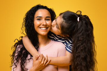 Wall Mural - Mothers Day Concept. Adorable Little Girl Embracing And Kissing Her Arab Mom, Portrait Of Happy Middle Eastern Mother And Daughter Bonding Together Over Yellow Studio Background, Copy Space