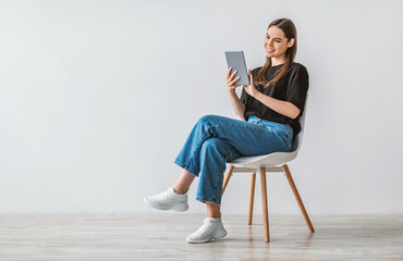 Poster - Full length portrait of cheerful young woman with digital tablet sitting on chair, working online against white studio wall, free space. Modern technologies and communications concept