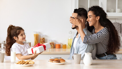 Wall Mural - Loving middle-eastern mother and daughter greeting their father with birthday, closing his eyes and giving big festive gift box while having breakfast together in kitchen. Family birthday celebration