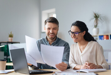 Poster - Cheerful male and female company workers looking through documents, collaborating, using laptop at modern office. Happy colleagues reading business papers, working on startup project together