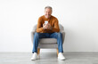 © Prostock-studio - Front portrait of smiling mature man using mobile phone, watching video sitting on armchair isolated on white studio background wall. Guy browsing internet, surfing web, free copy space