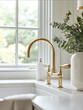 © inssaaet - Closeup of a brass faucet on a white sink in a farmhouse kitchen with a large window and neutral decor soft natural light vintage style simple elegance warm colors a