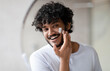 © Prostock-studio - Facial skincare. Indian man applying moisturizing cream on face, looking at his reflection in bathroom mirror at home, free space. Male beauty and skin care concept