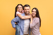 © Prostock-studio - A happy family poses against a yellow background. The father holds his daughter on his back while the mother stands close, creating a warm atmosphere of love and togetherness.
