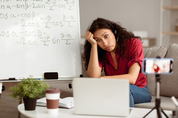 Aufkleber - Portrait of shocked and tired lady teacher in headset having virtual class, recording online lesson using smartphone on tripod, looking at laptop screen, sitting at table near whiteboard