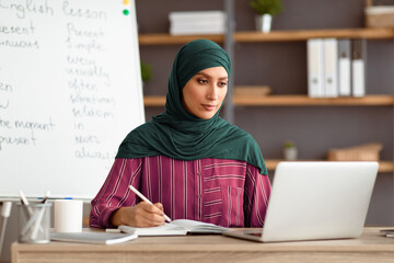 Aufkleber - Online Education. Lady muslim English teacher sitting at desk using laptop having video conference chat with students and class group. Woman wearing hijab looking at pc screen, writing in notebook