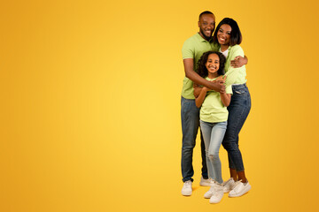 Wall Mural - Healthy family relationship. African american dad and mom embracing smiling daughter standing over yellow studio background, crop. Full length portrait