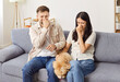 © Studio Romantic - Young family couple sitting on sofa with their maltipoo dog, suffering from bad smell. Displeased husband and wife holding their noses with their hands, looking at dirty pet with unhappy grimace.