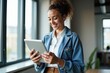 © velikiyzayats - Smiling young woman in casual denim jacket using tablet in bright, modern office setting, perfect for tech, business, or lifestyle marketing.