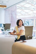 © BullRun - Chinese female employee with smartphone technology in hand sitting at desk in office looking away and smiling, prosperous Asian woman in smart casual outfit enjoying day in modern coworking space