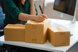 © Phushutter - An Asian young woman sits in a room, working on a computer and making calls or video calls, managing her small business and online sales, surrounded by cardboard boxes ready for shipping.