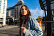 © we.bond.creations - A curious woman carrying a denim jacket and a backpack walking through the busty city and looking aside.