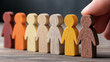 © Gabriele - Hand selecting a wooden figure from a row of diverse wooden figurines representing individuality and diversity, inclusion concept, wooden table background, equality and leadership selection idea