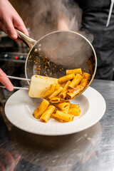 Wall Mural - A chef elegantly pours steaming rigatoni pasta onto a white plate, showcasing a delicious sauce. The kitchen ambiance adds a warm, inviting feel to the culinary experience.