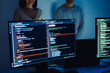 © weedezign - Close-up of computer screens displaying code, with developers collaborating in the background