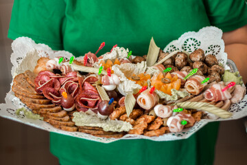 A green shirted individual holds a platter of appetizers featuring bacon, salami, skewered vegetables, meatballs, crackers, and garnishes.