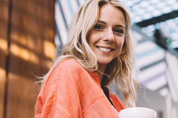 Half length portrait of cheerful blonde woman 20s in casual wear resting with aroma coffee outdoors, smiling attractive hipster girl looking at camera holding mug satisfied with free time recreation