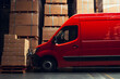 © elina - Red delivery van parked in a warehouse loading area surrounded by stacked cardboard boxes during the daytime