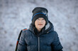 © Emvats - A smiling boy in a black winter jacket and hat lightly dusted with snow poses outdoors on a snowy day. A cheerful and warm expression in a cold setting.