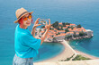 © EdNurg - A traveler woman stands at the viewpoint of Sveti Stefan island in Montenegro seacoast and taking photo on smartphone