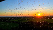 © Caroline - A windowpane adorned with glistening raindrops, framing a view of a vibrant green landscape bathed in the warm glow of a setting sun.