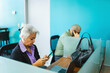 © STOCKEROcr - An older woman uses her mobile phone while working at a desk with a laptop, and a man in the background talks on the phone.
