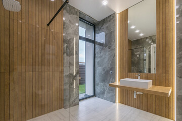 Bathroom with an exclusive design in a luxurious mansion. The room with a shower behind a glass partition is decorated with gray marble tiles and wooden panels.