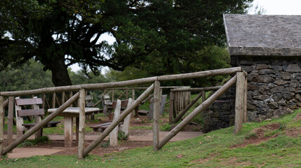 Wall Mural - A rustic picnic area with wooden tables, benches, and a wooden fence, set near a stone building and surrounded by lush trees. The scene exudes a peaceful, countryside atmosphere.