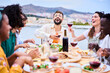 © CarlosBarquero - Joyful young adult friends holding red wine glass celebrating rooftop barbecue summer party. Happy millennial people laughing enjoying together with alcoholic beverages sitting at food table outdoor