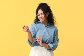 Wall Mural - Pretty young African-American woman buttoning up stylish blue shirt on yellow background