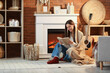 © Pixel-Shot - Young woman with warm plaid reading book near fireplace at home in evening