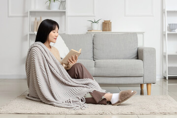 Wall Mural - Young Asian woman with grey blanket reading book on floor at home