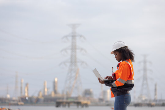 A female engineer wearing a white safety helmet and orange reflective jacket stands outdoors holding a clipboard, observing and analyzing a power plant facility with electrical towers in the backgroun