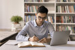 © fizkes - Doing homework. Focused male teenager in eyewear write literature review at college university library work on research project for student science contest study paper books online sources on notebook