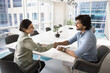 © fizkes - Happy successful Indian business partners shaking hands in collaboration room, discussing teamwork, career, leadership at large table. Female business leader giving greeting handshake to colleague