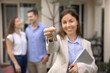 © fizkes - Happy successful female real estate agent offering key, showing bunch at camera, posing for professional realtor portrait with rental accommodation and couple of renters, buyers in blurred background