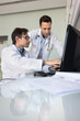 © fizkes - Team of two male doctors reviewing clinical medical case at computer together, looking and pointing at monitor at workplace table, talking, discussing examination report, diagnosis. Vertical shot