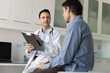 © fizkes - Caring young male doctor meeting with patient in clinic examination room, asking about symptoms, making records in paper documents, writing notes, talking to patient. Low angle candid shot