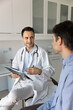 © fizkes - Positive general practitioner man talking to patient on appointment in clinic office, holding pad with medical records, speaking, discussing complaints, recommendation, explaining treatment